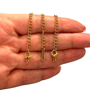May include: Three gold-colored chain extenders with lobster claw clasps. The extenders are made of a delicate chain link design. The clasps are attached to the chain with a small loop. The extenders are shown against a white background.