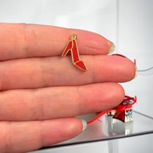 Può includere: Un piccolo ciondolo a forma di scarpa con tacco alto rosso con un contorno dorato. Il ciondolo è tenuto dalle dita di una persona. Il ciondolo è una versione in miniatura di una scarpa con tacco alto.