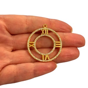 May include: Gold-coloured pendant with a circular design featuring Roman numerals and a smaller inner circle. The outer ring is encrusted with small, clear stones. The pendant has a loop at the top for hanging.