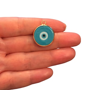 May include: A circular pendant featuring a stylized evil eye design with a central black pupil, a white iris, and a light blue outer ring, all encased in a thin gold-toned metal frame with a small circular bail at the top for attachment.