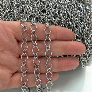 May include: A continuous spool of silver-toned metal chain featuring elongated, diamond-shaped links connected by smaller circular jump rings. The chain has a polished, reflective finish and is presented in a dense, coiled arrangement.