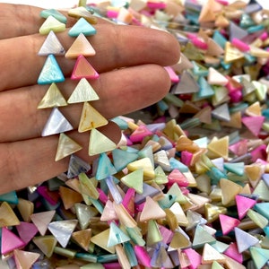 May include: A hand holding a strand of colourful, triangular shell beads. The beads are in shades of pink, blue, yellow, and white. A pile of similar beads is in the background.