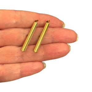 May include: Two gold-coloured metal bar components, each featuring a smooth, cylindrical, tube-like shape with a small, flat, circular attachment point at the top end. These metallic pieces have a polished, reflective finish and a slender, elongated form.
