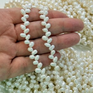 May include: A collection of small, off-white, teardrop-shaped freshwater pearls. The pearls feature a smooth, lustrous surface with natural variations in shape and size, arranged in loose strands and a large pile of individual loose beads.
