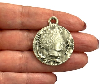 May include: Silver tone pendant charm featuring a profile of a woman with curly hair surrounded by three dolphins. The pendant is round and has a loop at the top for attaching to a chain or cord.