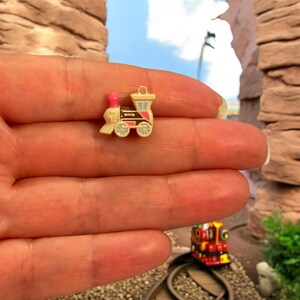 May include: A small train charm with a pink chimney, cream-coloured body, and red and black details. The train has light blue wheels and a gold-coloured loop for attaching. The charm is held in a hand. The miniature train is a decorative item.