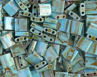 May include: A close-up of a pile of turquoise blue and brown square glass beads. The beads have a mottled, iridescent finish.