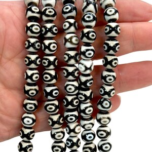 May include: Strands of black and white patterned beads. The beads have a circular design with a central eye-like pattern. The beads are strung together, creating a necklace or bracelet.