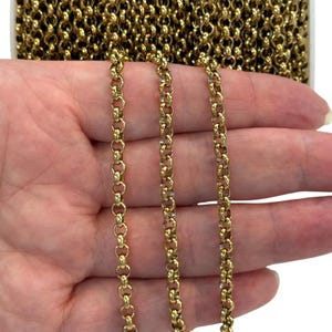Può includere: Primo piano di maglie di catena color oro. La catena è composta da piccole maglie rotonde collegate tra loro. La catena è tenuta in una mano, con un rotolo di catena visibile sullo sfondo. La catena è di un colore oro brillante e lucente.