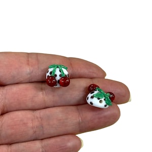 May include: Two white ceramic buttons with black polka dots and red cherries on top. The cherries have green stems and leaves.