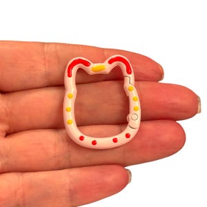 May include: A pink metal clasp in the shape of a cat's face, with red and yellow accents. The clasp has a hinged opening and is decorated with red and yellow dots. The clasp is held in a hand.