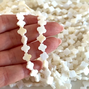 May include: A strand of white shell beads in the shape of four-leaf clovers. The beads are strung on a white thread and are displayed in a hand.