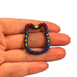 May include: A blue carabiner clip with red and yellow accents. The clip has a rounded shape with a hinged opening. Yellow and red dots decorate the clip's surface. The clip is held in a hand.