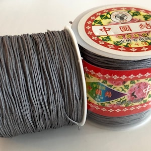 May include: Two spools of gray elastic cord, each with a white plastic spool and a red, white, and green label with Chinese characters. The label on the spool on the right says "中国 结" which translates to "Chinese knot".