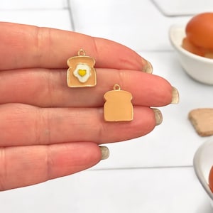 Op de afbeelding: Twee goudkleurige bedels in de vorm van toast. Een bedel heeft een wit en geel geëmailleerd ei met een geel hart in de dooier. De andere bedel is effen.
