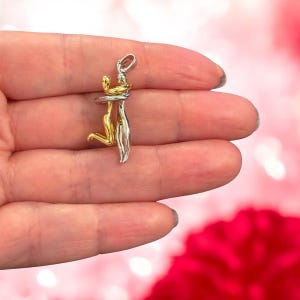 May include: A pendant depicting two figures in an embrace. One figure is gold-coloured, the other silver-coloured. The pendant hangs from a small loop. The background is a soft pink with a blurred red floral element.