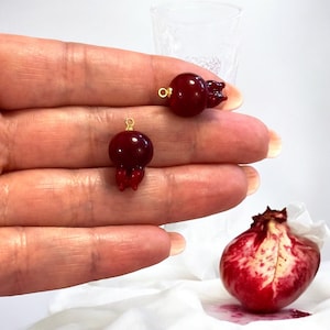 May include: Two red glass pomegranate charms with gold jump rings. The charms are on a hand, and a whole pomegranate is in the background.