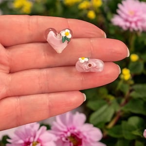 Puede incluir: Dos cuentas de vidrio rosa con flores blancas y hojas verdes. Las cuentas son de forma de corazón y redondas.