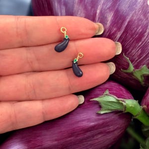 May include: Two gold-coloured eggplant charms with green accents. The charms are small and are held in a hand.