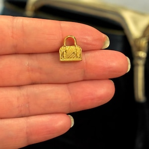 May include: A small, gold-colored handbag charm with a textured surface and a curved handle. The charm is rectangular with a detailed design, likely for jewelry or accessories.