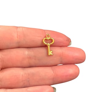 Pode incluir: Um pequeno pingente de chave dourada com um topo em forma de coração e um detalhe de chave. O pingente é projetado para fazer joias ou artesanato. A chave é segurada entre os dedos em um fundo branco.