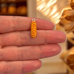 May include: A miniature baguette charm with a silver-coloured loop. The charm is yellow with orange stripes and small black dots, resembling a loaf of bread. This food-themed charm is suitable for jewellery making.