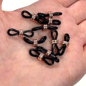May include: A close-up of eight black rubber eyeglass holders with rose gold accents. The holders are shaped like a figure eight and have a small loop on each end.