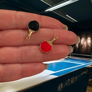 May include: Two gold-coloured table tennis paddle charms, one with a red surface and one with a black surface.