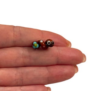Op de afbeelding: Een handgemaakte glazen kraal in de vorm van een rups. De kraal is bruin met witte stippen en heeft een bloem met turquoise bloemblaadjes en een paar wiebelogen. De kraal is ongeveer 2,5 cm lang.