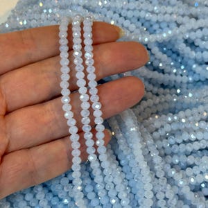 Könnte beinhalten: Nahaufnahme von mehreren Strängen hellblauer, facettierter Perlen. Die Perlen sind klein, rund und haben eine glitzernde, schillernde Oberfläche. Sie sind aufgefädelt und erzeugen ein zartes Aussehen. Die Perlen haben eine helle, eisblaue Farbe.