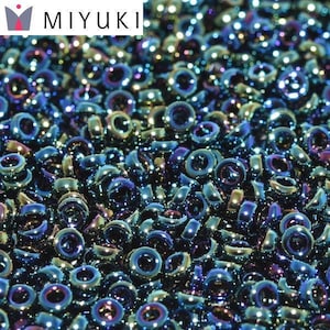 Op de afbeelding: Een close-up van een grote hoeveelheid kleine, iriserende blauwe en groene glazen kralen. De kralen zijn rond met een gat in het midden. De kralen zijn in een willekeurig patroon gerangschikt. De kralen zijn allemaal even groot. De kralen zijn glanzend en reflecterend. De kralen zijn perfect voor het maken van sieraden.