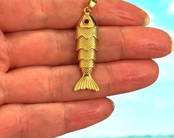 May include: A gold-colored fish pendant with a textured body and a small red accent on the eye. The fish has a detailed tail and is suspended from a small loop. The pendant is likely made of metal and is designed to be worn as jewelry.