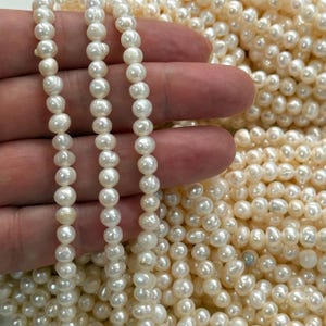 Peut inclure: Une collection de petites perles d'eau douce rondes de couleur blanc cassé. Trois brins individuels sont tenus verticalement, mettant en valeur la taille uniforme et la texture de surface légèrement irrégulière et lustrée de chaque perle.