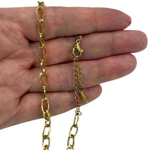 Könnte beinhalten: Goldfarbene Kette mit länglichen ovalen Gliedern. Die Halskette hat einen Karabinerverschluss und eine verstellbare Kette. Die Kette wird in einer Hand vor weißem Hintergrund gehalten. Die Halskette ist ein Schmuckstück.