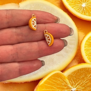 Op de afbeelding: Twee goudkleurige bedels in de vorm van sinaasappelschijfjes. De bedels bevinden zich op een hand met een achtergrond van sinaasappelschijfjes.