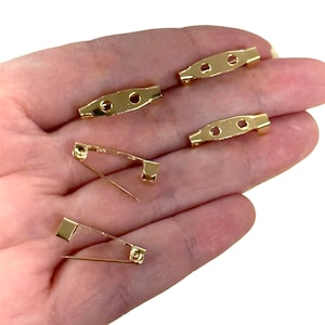 May include: Five gold-tone metal pin backs for attaching brooches, pins, or other jewelry to clothing. The pin backs are in various sizes and shapes.