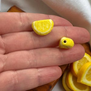 Op de afbeelding: Twee gele keramische kralen in de vorm van citroenpartjes met witte accenten. Eén kraal is een halve cirkel, de andere is een kleine kubus met een gat. De kralen liggen op een hand, met meer citroenpartjes op de achtergrond.