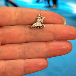 May include: A miniature silver charm depicting an ice skate. The skate features a white boot with a pink wing and ornate detailing. The charm is designed with a loop at the top for attachment to a bracelet or necklace, perfect for jewellery making.