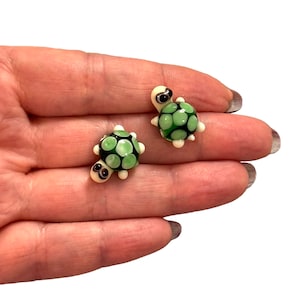 May include: Two small green glass turtle figurines with white accents and black eyes. The turtles are sitting on a hand.