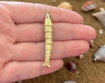 May include: A gold-colored fish pendant with a textured, segmented body and a small loop at the top for a chain. The pendant is held in a hand, with seashells and sand in the background. The fish pendant is approximately 2.5 inches long.