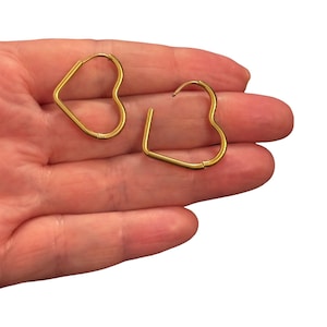 May include: A pair of gold-toned heart-shaped hoop earrings. Each earring features a thin, smooth metal wire bent into a heart outline with a hinged closure mechanism at the top, showcasing a simple and clean design for these jewellery pieces.