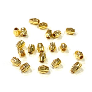 May include: A collection of gold-colored metal beads with a faceted design. The beads are scattered on a white surface.