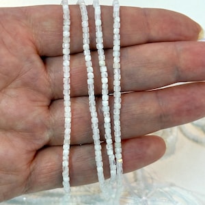 May include: Close-up of several strands of small, white, square-cut beads. The beads are strung together, creating a delicate and shimmering effect. The beads are held in a hand, showcasing their size and texture. Ideal for jewelry making, the beads have a simple elegance.