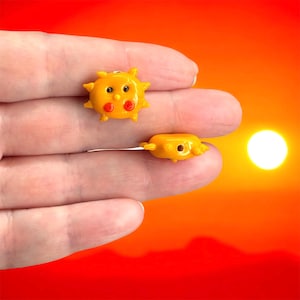 May include: Two small, yellow sun-shaped charms with black eyes and red cheeks. One charm is upright, the other is lying down. The charms are held in a hand against a vibrant orange and yellow background.