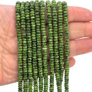 May include: Strands of green gemstone beads with black speckles. The beads are small, round, and uniform in size, strung together to create a flowing, textured appearance. The beads are held in a hand, showcasing their color and detail.