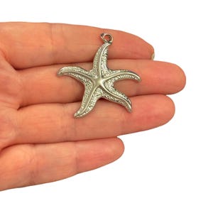 May include: A silver-toned metal starfish pendant featuring a textured, granular surface on its five arms and a smooth, raised central star shape. The piece includes a small circular jump ring at the top for attachment to a chain or cord.