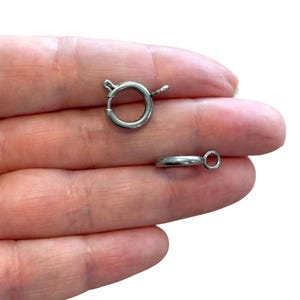 Puede incluir: Dos cierres de anillo de resorte plateados. Uno está abierto, mostrando el anillo circular y el mecanismo de resorte. El otro es una pequeña barra curva con un pequeño anillo adjunto. Estos cierres se utilizan para la fabricación de joyas.