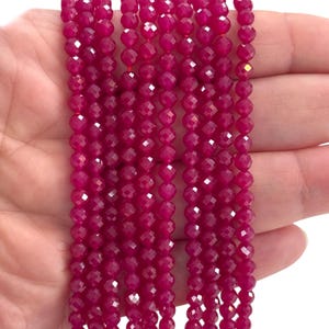 Puede incluir: Primer plano de varios hilos de cuentas facetadas de color magenta intenso. Las pequeñas cuentas redondas están ensartadas, creando un efecto brillante. Las cuentas se sostienen en una mano, mostrando su color y textura. Ideal para la fabricación de joyas.