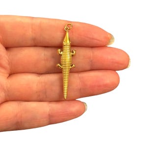 Pode incluir: Um pingente de metal em tom dourado com formato de jacaré ou crocodilo, apresentando uma superfície texturizada e escamosa em todo o corpo e membros, com um pequeno anel circular fixado no topo para pendurar em uma corrente ou cordão.