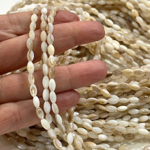Op de afbeelding: Close-up van strengen ovaalvormige, gebroken witte kralen met subtiele bruine marmering. De kralen zijn aan elkaar geregen, waardoor een delicate textuur ontstaat. De achtergrond toont een grotere stapel vergelijkbare kralen.